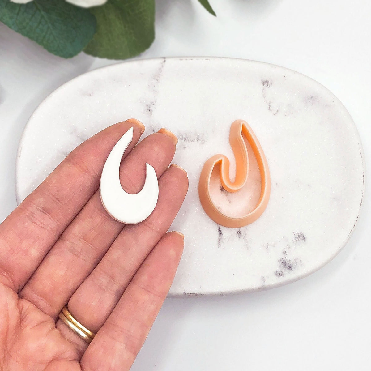 A hand holds a small white organic droop-shaped cut out next to a peach-colored clay cutter on a white marble dish.