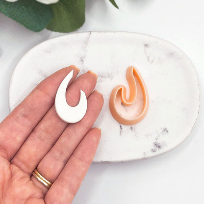 A hand holds a small white organic droop-shaped cut out next to a peach-colored clay cutter on a white marble dish.