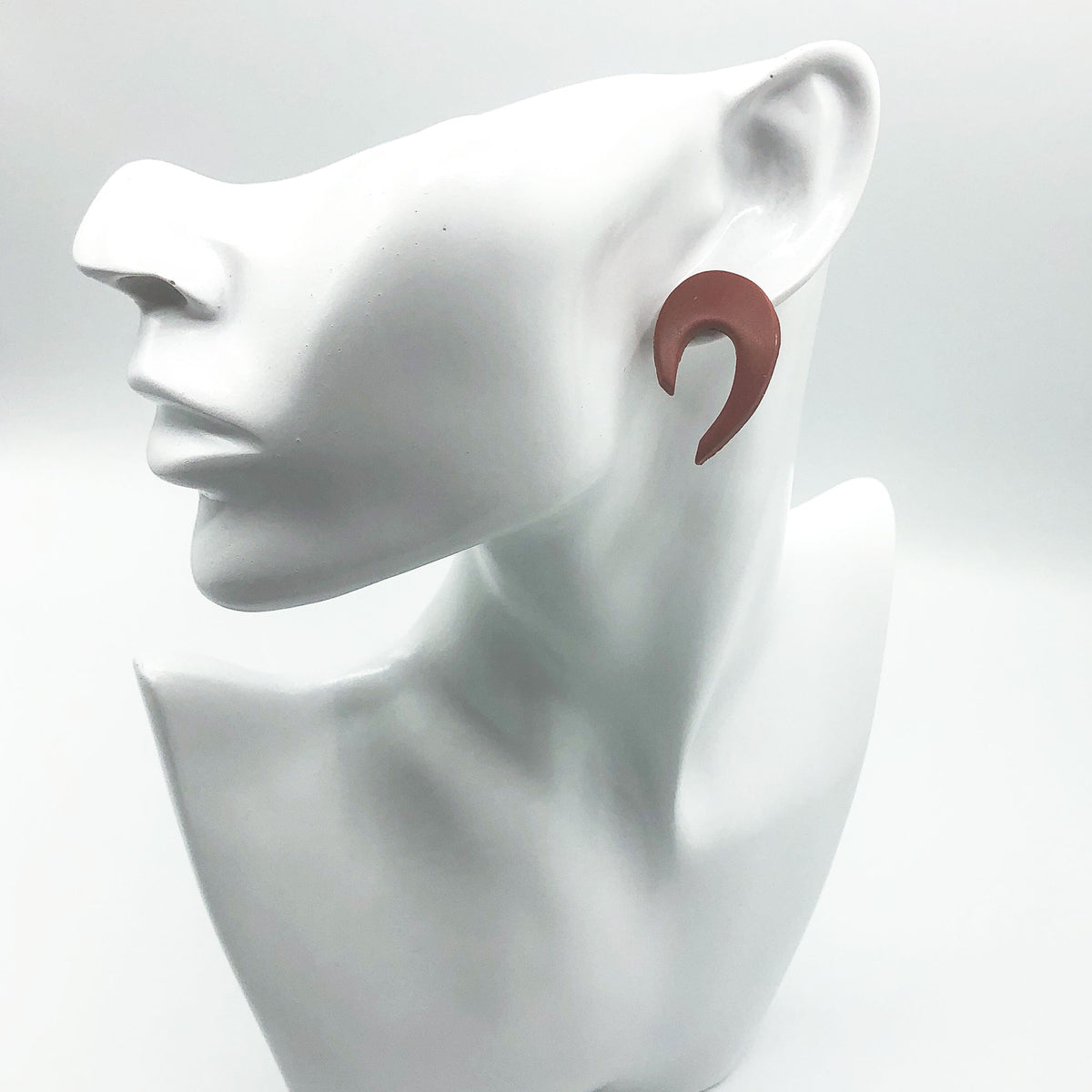 A single matte terracotta-colored curved hoop earring displayed on a white ceramic female bust against a light gray background.