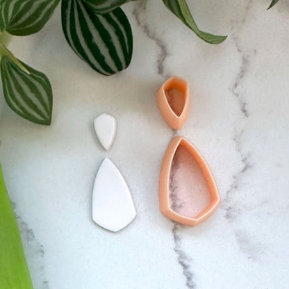 Shield teardrop-shaped clay charm in white and peach-orange clay cutter arranged on a white marble surface with green plant leaves in the background.