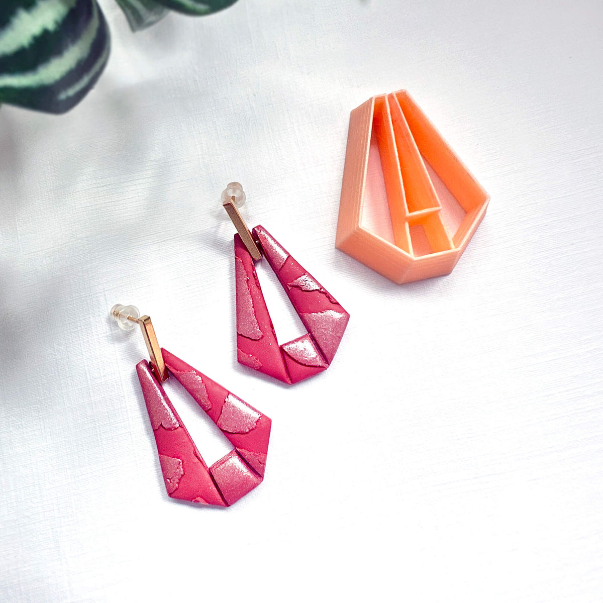 A pair of geometric dangle earrings in a bright pink and magenta patterned polymer clay with gold-tone posts, displayed next to an orange geometric clay cutter on a white textured background.