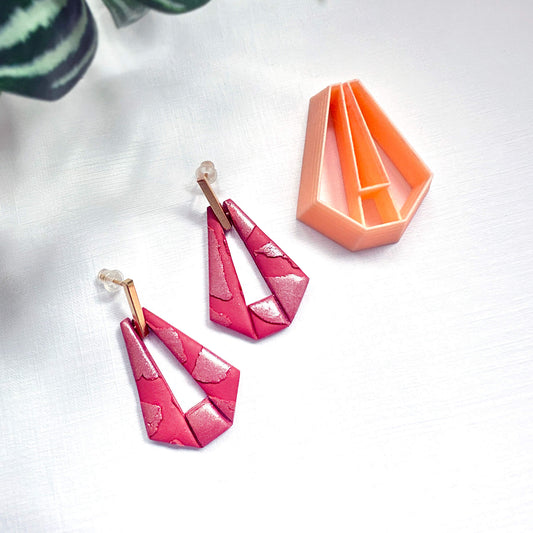 A pair of geometric dangle earrings in a bright pink and magenta patterned polymer clay with gold-tone posts, displayed next to an orange geometric clay cutter on a white textured background.