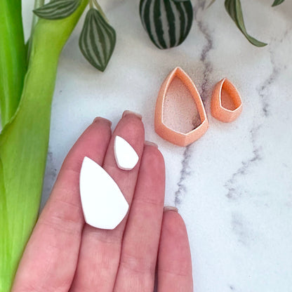 Shield teardrop-shaped clay cut outs in white and a peach-pink clay cutter are displayed on a white marble surface next to a person's hand for scale, with green leaves in the background.