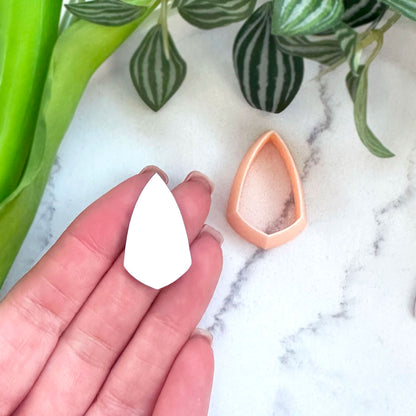 A hand holds a white shield teardrop-shaped clay cutout next to a peach-colored clay cutter on a white marble surface with green plants in the background.
