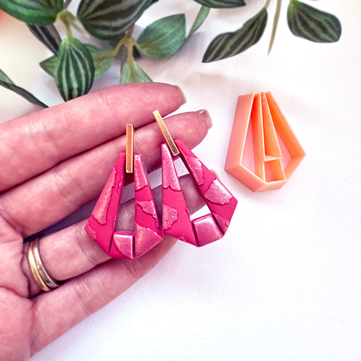 A pair of bright pink geometric dangle earrings with gold-tone posts, held in a person's hand next to an orange clay cutter, on a white background with green leaves.