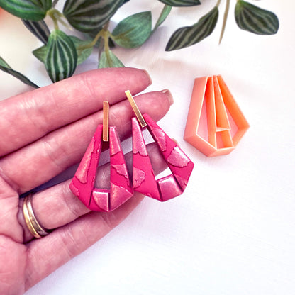 A pair of bright pink geometric dangle earrings with gold-tone posts, held in a person's hand next to an orange clay cutter, on a white background with green leaves.
