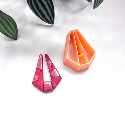 A small orange plastic geometric cookie clay cutter and a matching pink glittery cutout-shaped polymer clay piece on a white surface with blurred green leaves in the background.