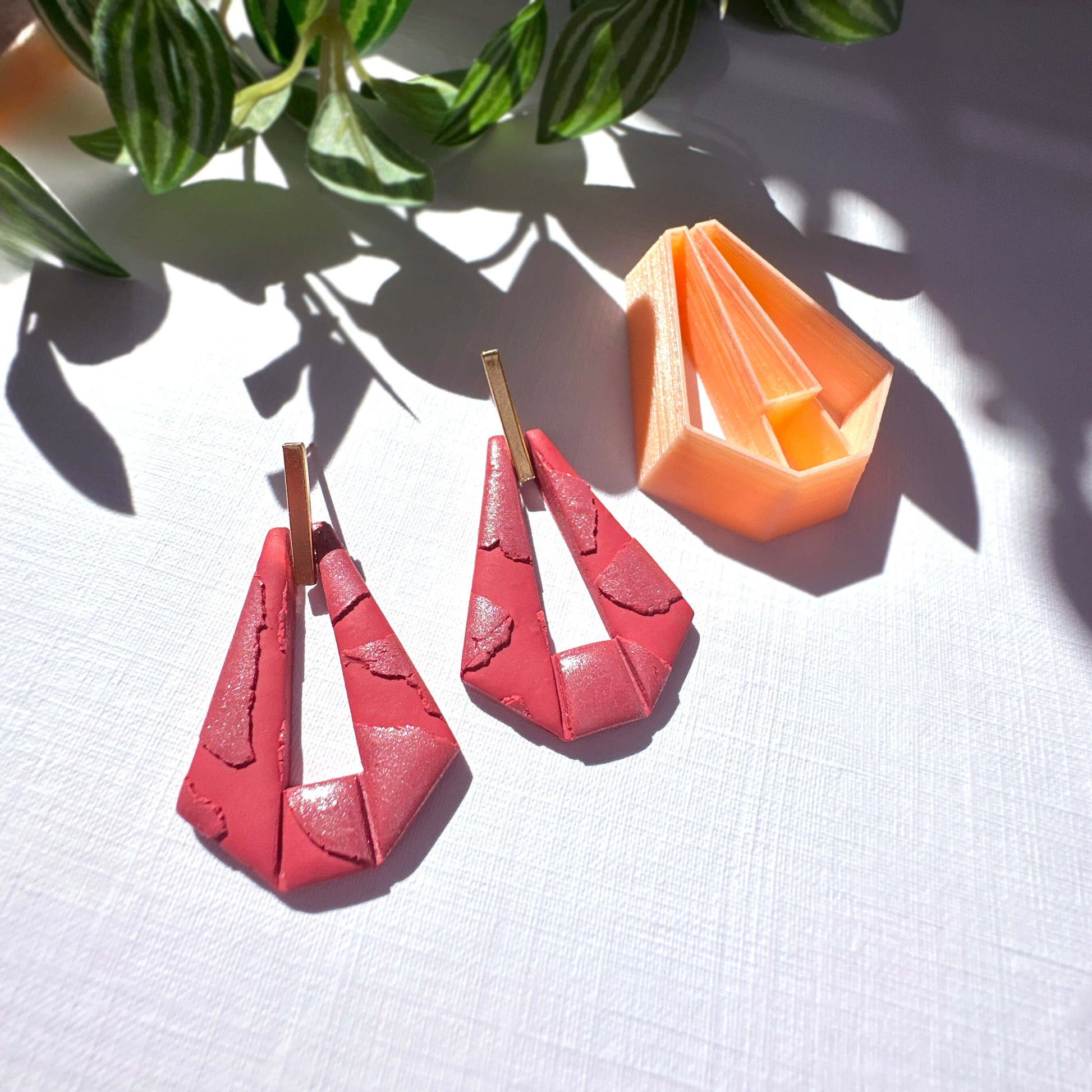 A pair of red geometric drop earrings with gold-tone posts, displayed on a white textured surface with leafy plant leaves casting shadows. An orange geometric clay cutter is visible in the background.