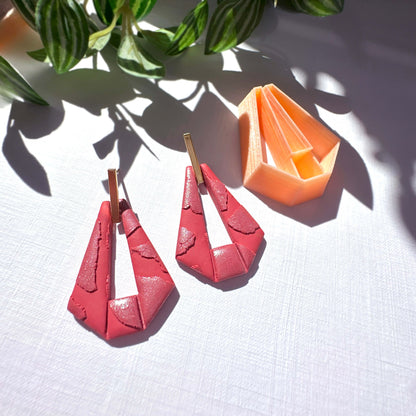 A pair of red geometric drop earrings with gold-tone posts, displayed on a white textured surface with leafy plant leaves casting shadows. An orange geometric clay cutter is visible in the background.