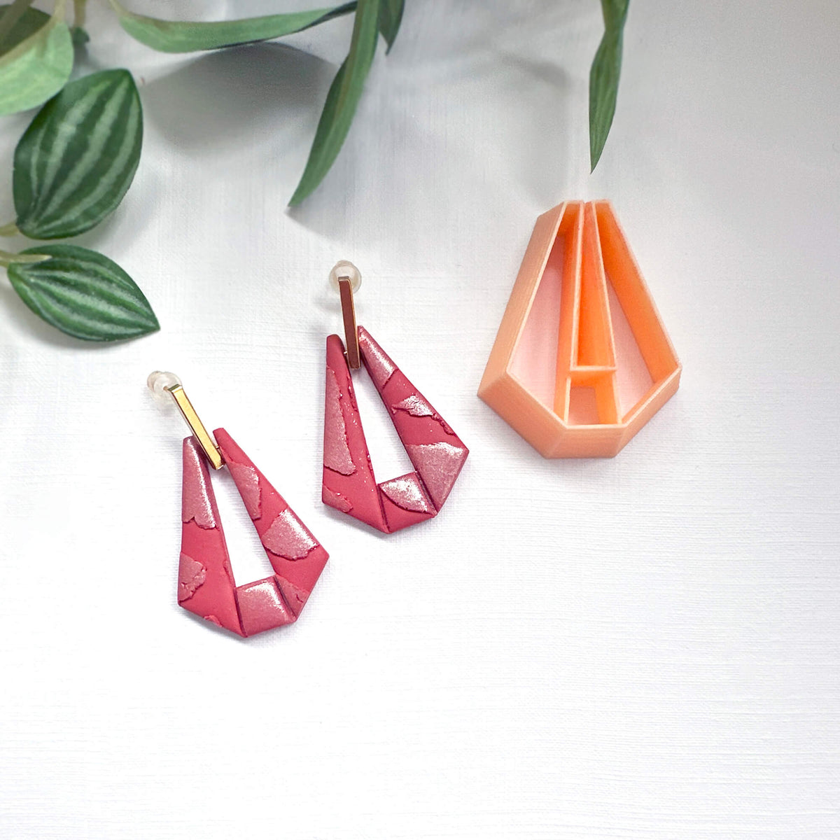 A pair of handmade geometric dangle earrings in a glossy pink and red marbled pattern with gold posts, displayed next to a peach-colored clay cutter on a white background with green leaves.