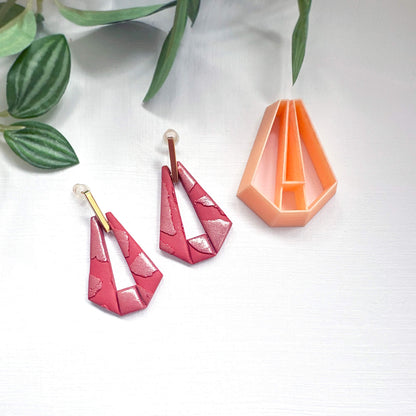 A pair of handmade geometric dangle earrings in a glossy pink and red marbled pattern with gold posts, displayed next to a peach-colored clay cutter on a white background with green leaves.