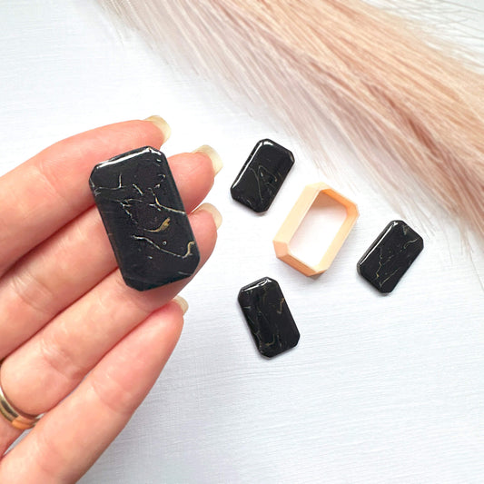 A hand holds a rectangular black polymer clay faceted rectangle shape with gold veining, while four matching black stone pieces and one peach-colored clay cutter are arranged on a light surface 
