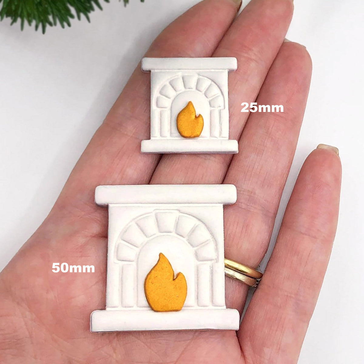 Two small white fireplace-shaped pooymer clay cut outs with yellow-orange flames are shown resting on a person's hand, with size labels reading 25mm and 50mm. The hand is against a clean white background with a small green plant visible in the top corner.