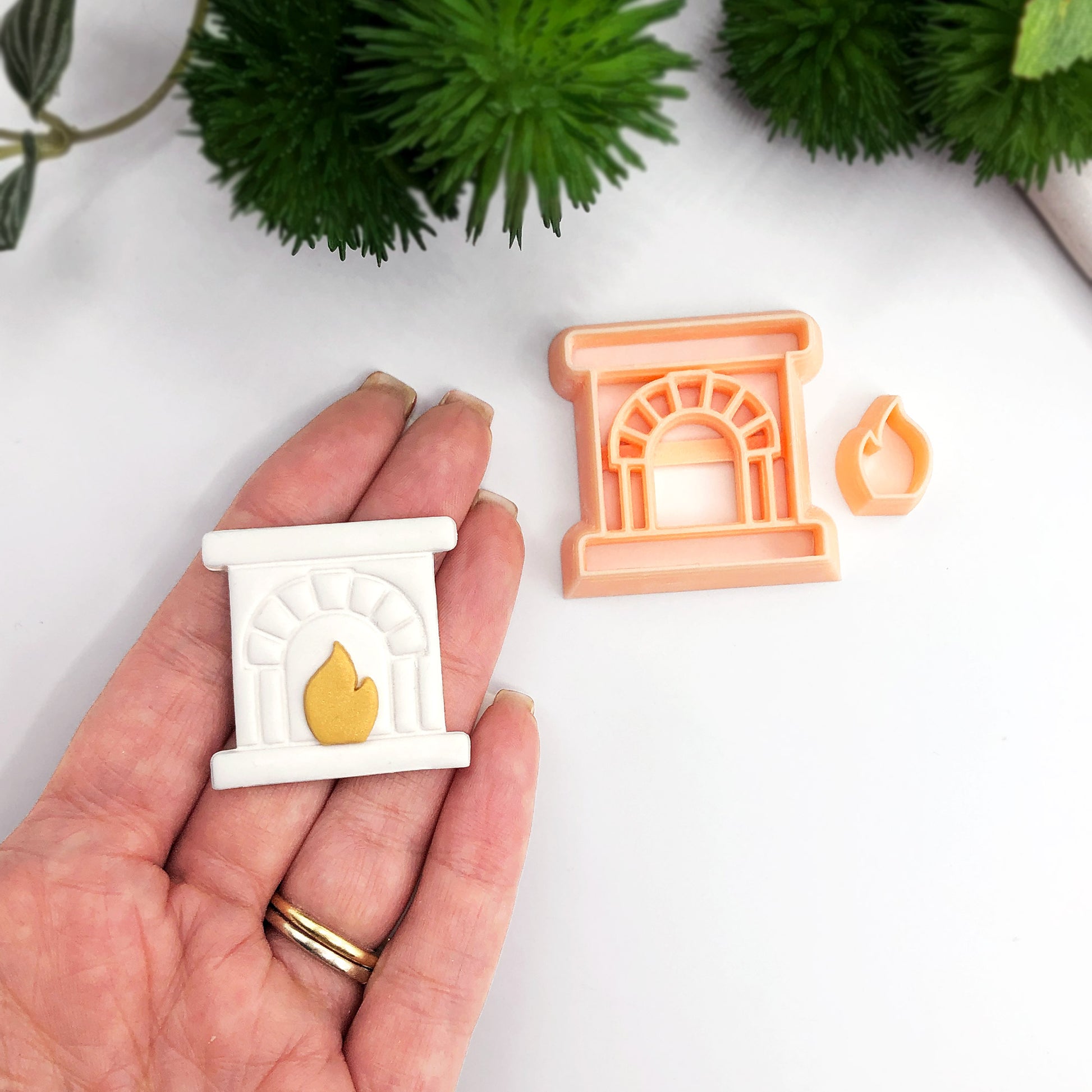 A hand holds a miniature white fireplace-shaped polymer clay cut out with a gold flame detail, next to a peach-colored clay cutter with a small matching flame piece, on a white surface with green plants in the background.