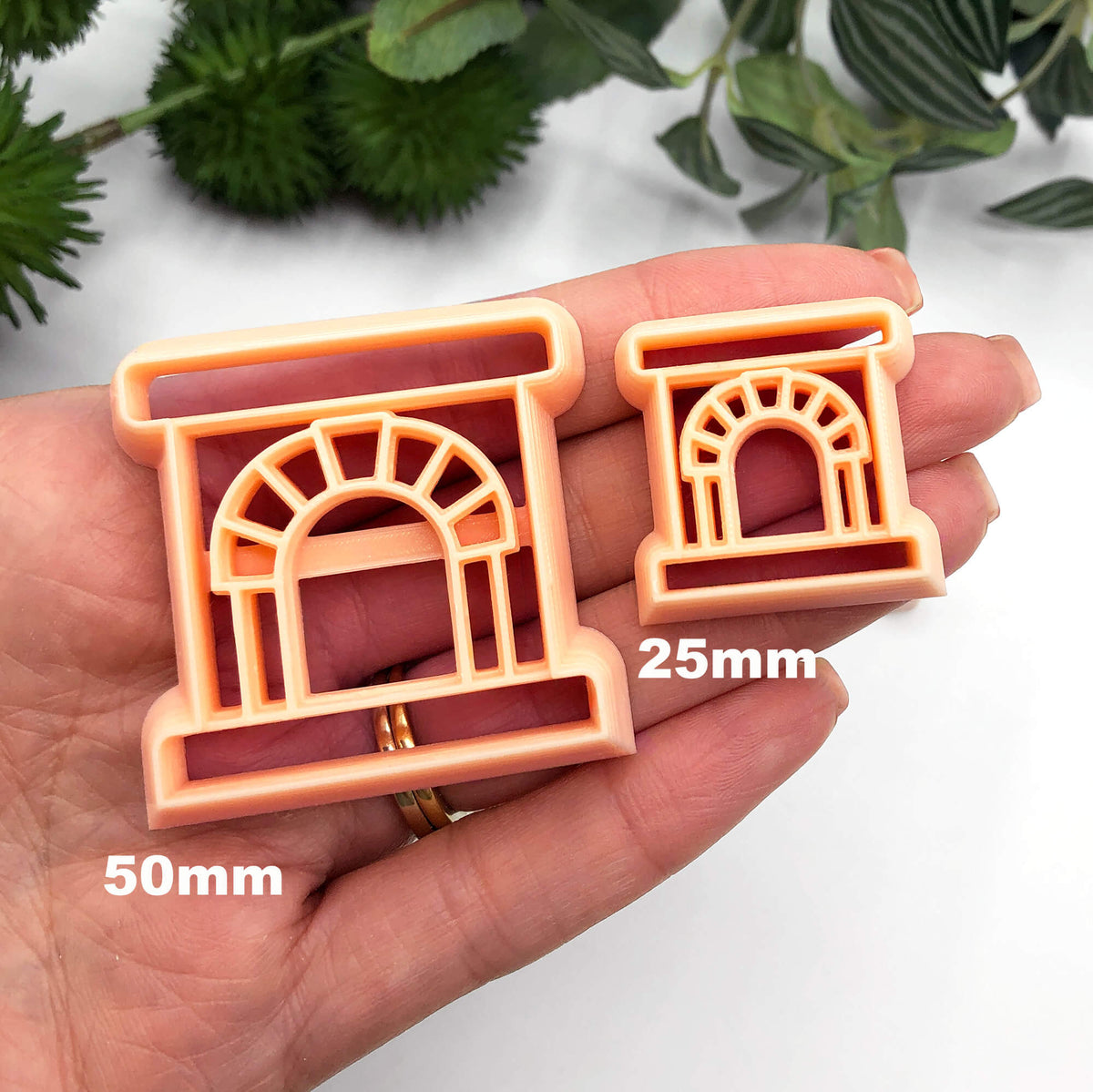 Two peach-colored plastic clay cookie cutters shaped like fireplaces, one 25mm, one 50mm held in a person's hand against a white background with blurred green foliage.