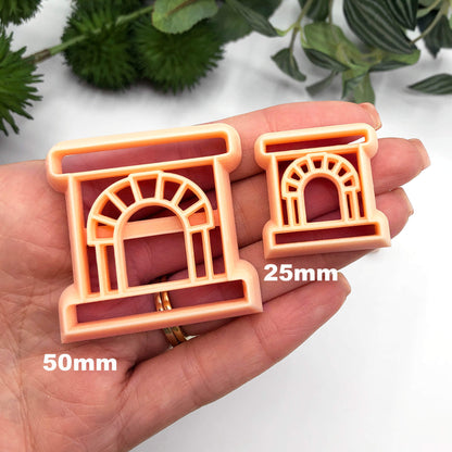 Two peach-colored plastic clay cookie cutters shaped like fireplaces, one 25mm, one 50mm held in a person's hand against a white background with blurred green foliage.
