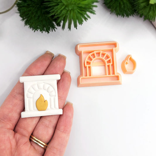 A hand holds a miniature white fireplace-shaped polymer clay cut out with a gold flame detail, next to a peach-colored clay cutter with a small matching flame piece, on a white surface with green plants in the background.