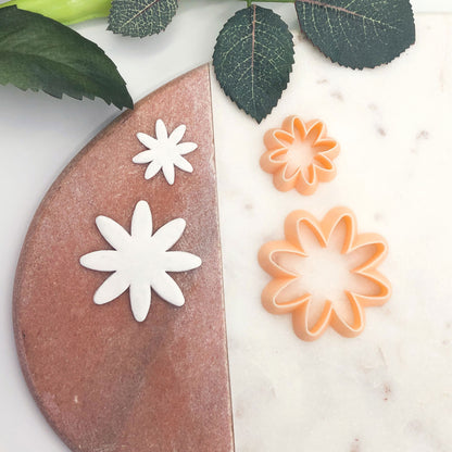Two white clay cut outs and two peach flower-shaped cookie cutters displayed on a split white marble and terracotta background with green leaves.