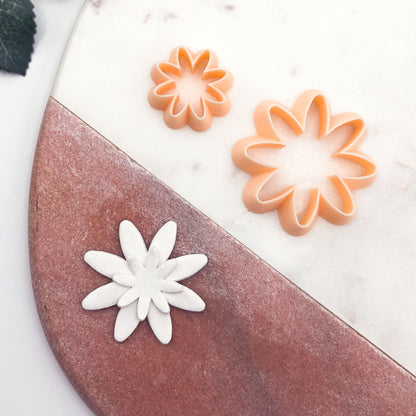 Two flower-shaped cookie cutters, one peach and one white, displayed on a split white and terracotta surface.
