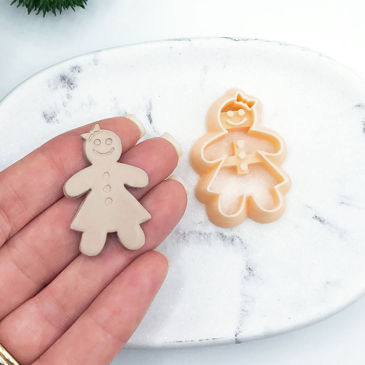 A hand holds a small gingerbread person clay cutout, with a matching gingerbread person cookie clay cutter resting on a white marble surface in the background.
