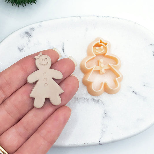 A hand holds a small gingerbread person clay cutout, with a matching gingerbread person cookie clay cutter resting on a white marble surface in the background.