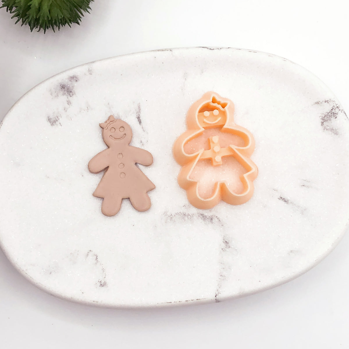 A gingerbread person cookie clay cutter in peach and a matching gingerbread person cutout in tan, displayed on a white marble tray.