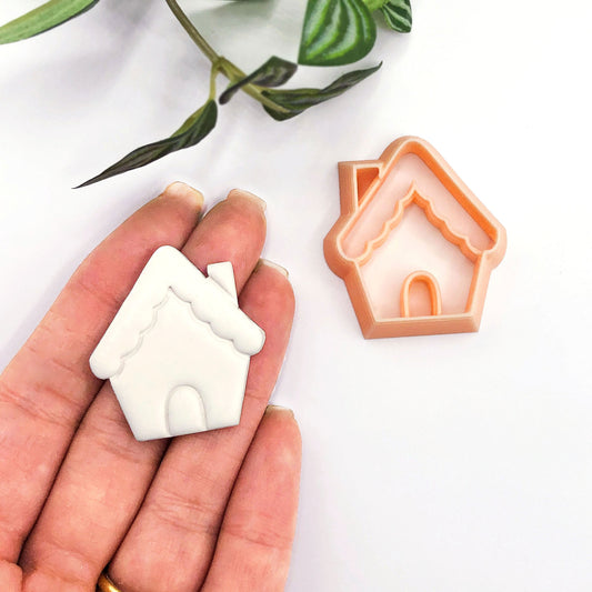 A small gingerbread house-shaped white item held in a person's hand next to an orange house-shaped cookie cutter or mold on a clean white background with a leafy stem at the top.