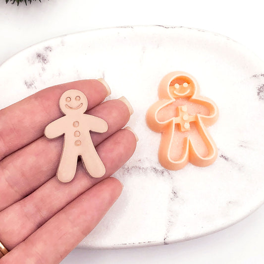 A hand holds a small gingerbread man-shaped clay cutout next to a gingerbread person-shaped cookie clay cutter on a white marble plate.