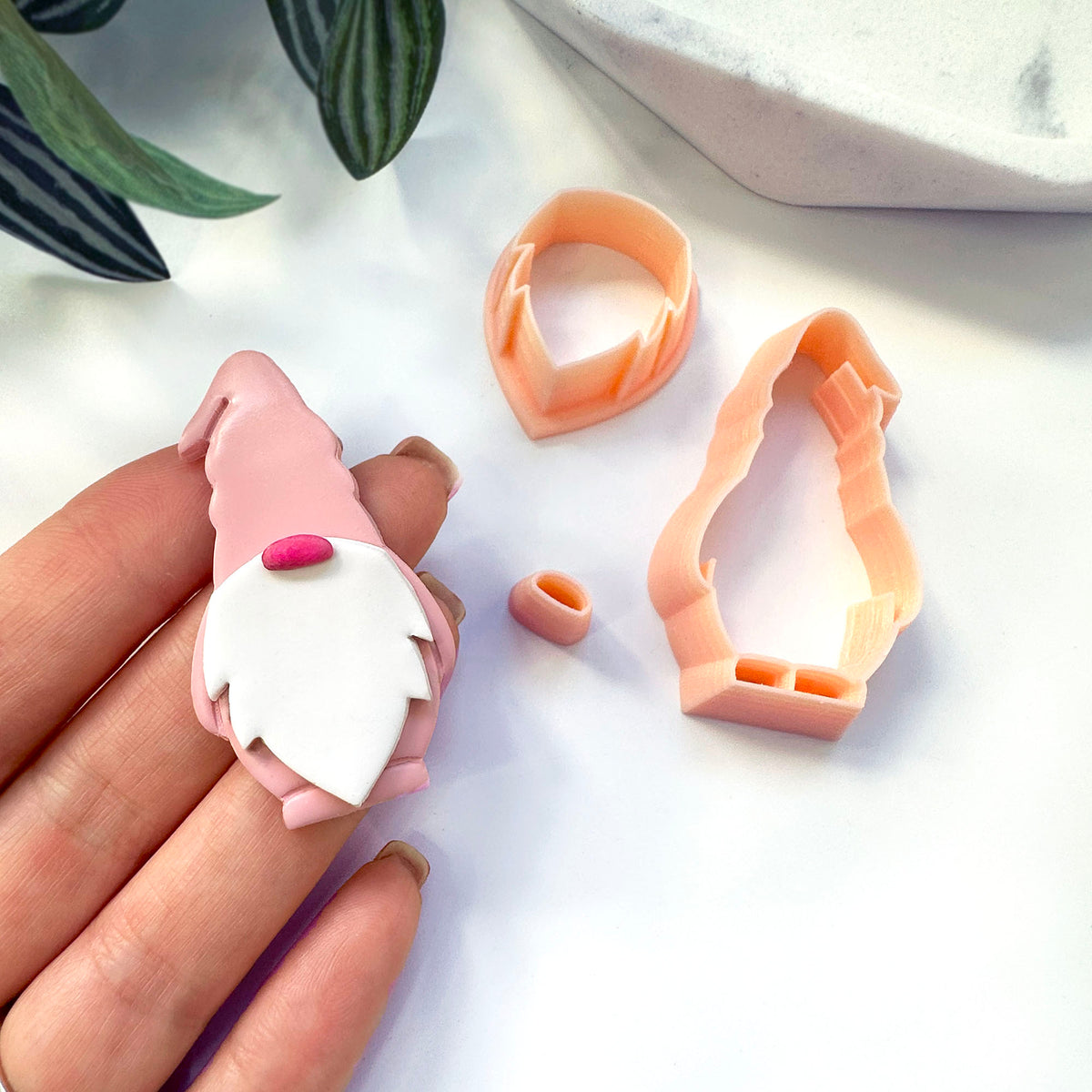 A hand holds a small pink gnome polymer clay figurine with a white beard and pink hat, next to peach-colored clay cutters and a small matching piece on a white surface with a plant leaf in the background.