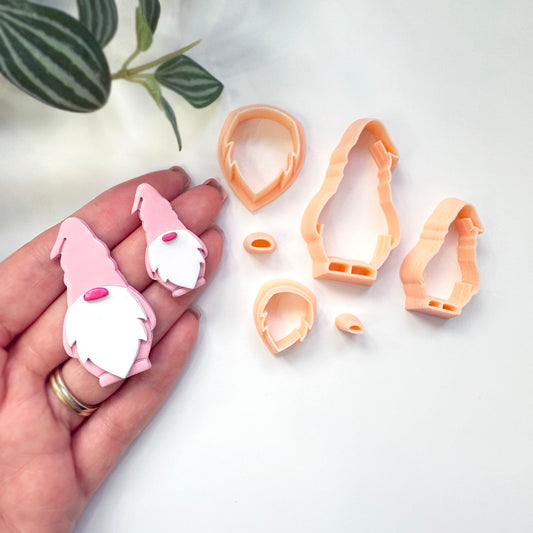 A hand holds two small gnome-shaped polymer clay cutouts next to several peach-colored plastic cookie cutters and cutters templates on a white surface with a green plant leaf in the corner.