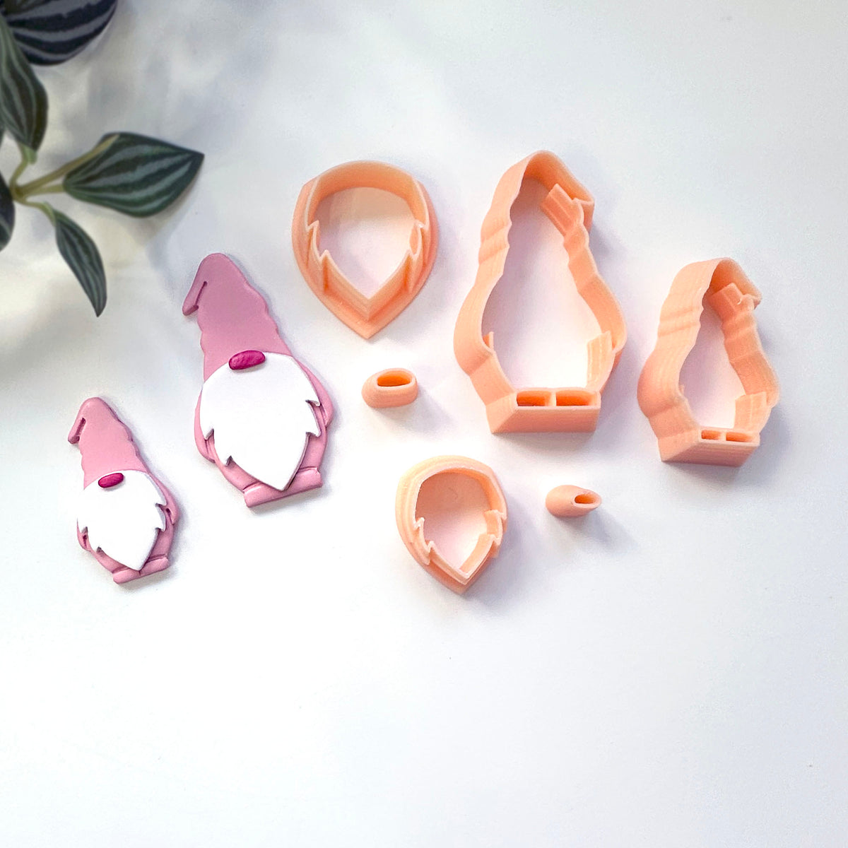 A set of gnome-shaped clay cookie cutters and matching gnome cutouts arranged on a white surface, with a small plant leaf visible in the top-left corner.y 