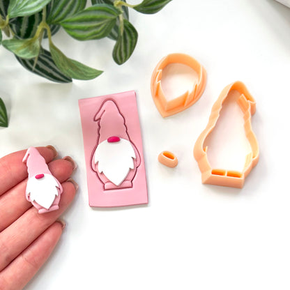 A hand holds a small gnome-shaped clay piece next to a pink cut out with peach-colored plastic cutter pieces arranged on a white surface. A green plant leaf is visible in the top left corner.