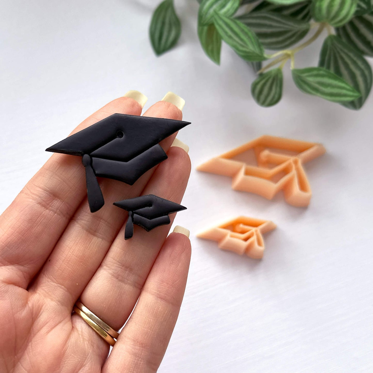 Black graduation cap cookie cutters held by a hand with green leaves in the background - polymer clay cutter