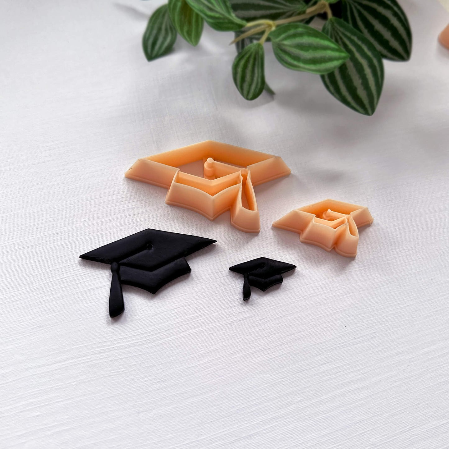 Graduation-themed cut-outs of black caps and the polymer clay cutters on a white surface with green leaves in the background