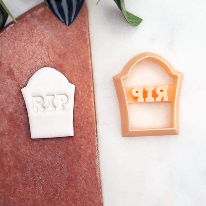 A Halloween-themed RIP tombstone cookie cutter shown next to a white clay tombstone with the letters 'RIP' cut out, displayed on a split terracotta and white marble background.