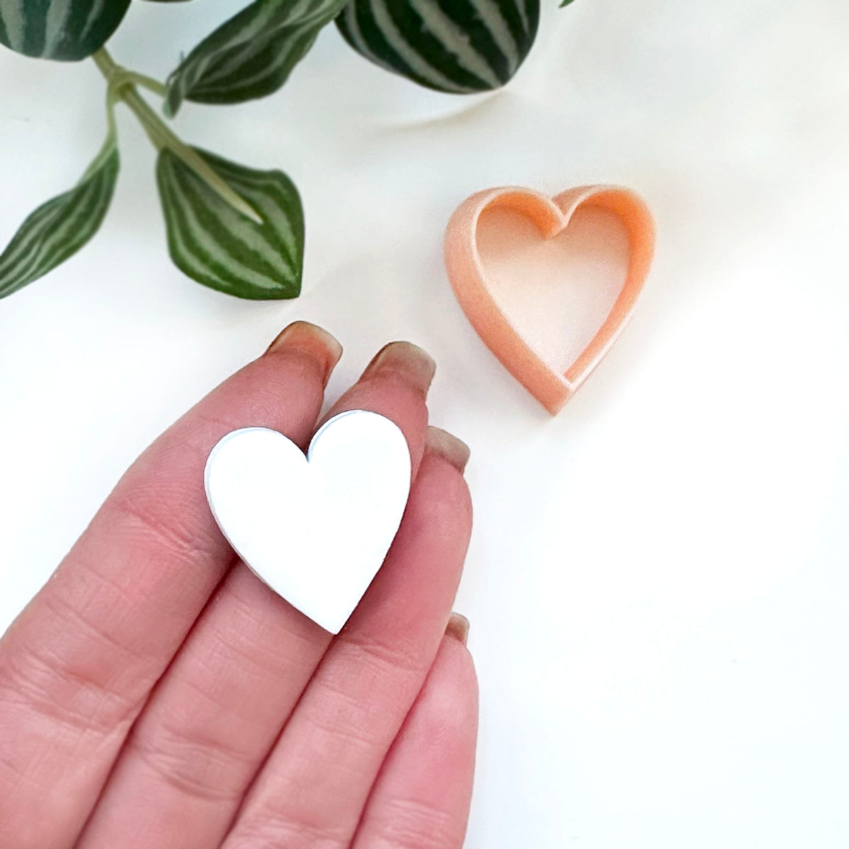 A hand holds a small white heart-shaped cut out while another peach-colored heart-shaped cutter sits on a white surface, with green leaves in the upper left corner.