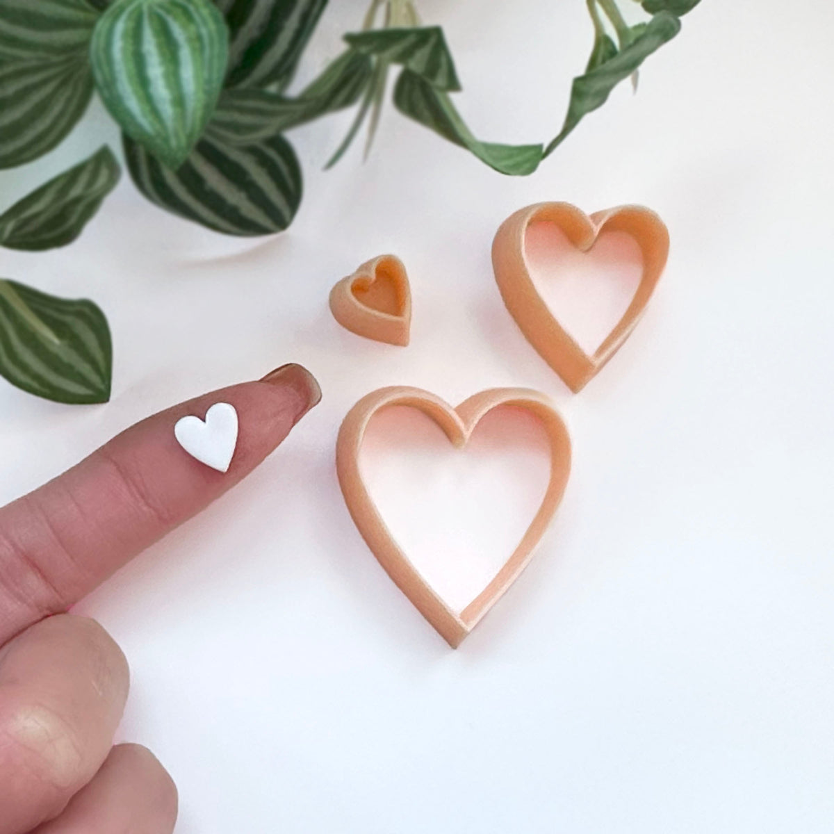 Three heart-shaped cookie clay cutters in a light peach-beige color are displayed on a white surface with green plant leaves in the background. A person's finger is visible on the left holding a small white heart-shaped cutout.