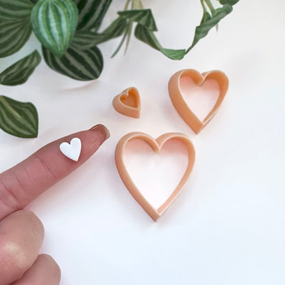 Three heart-shaped cookie clay cutters in a light peach-beige color are displayed on a white surface with green plant leaves in the background. A person's finger is visible on the left holding a small white heart-shaped cutout.