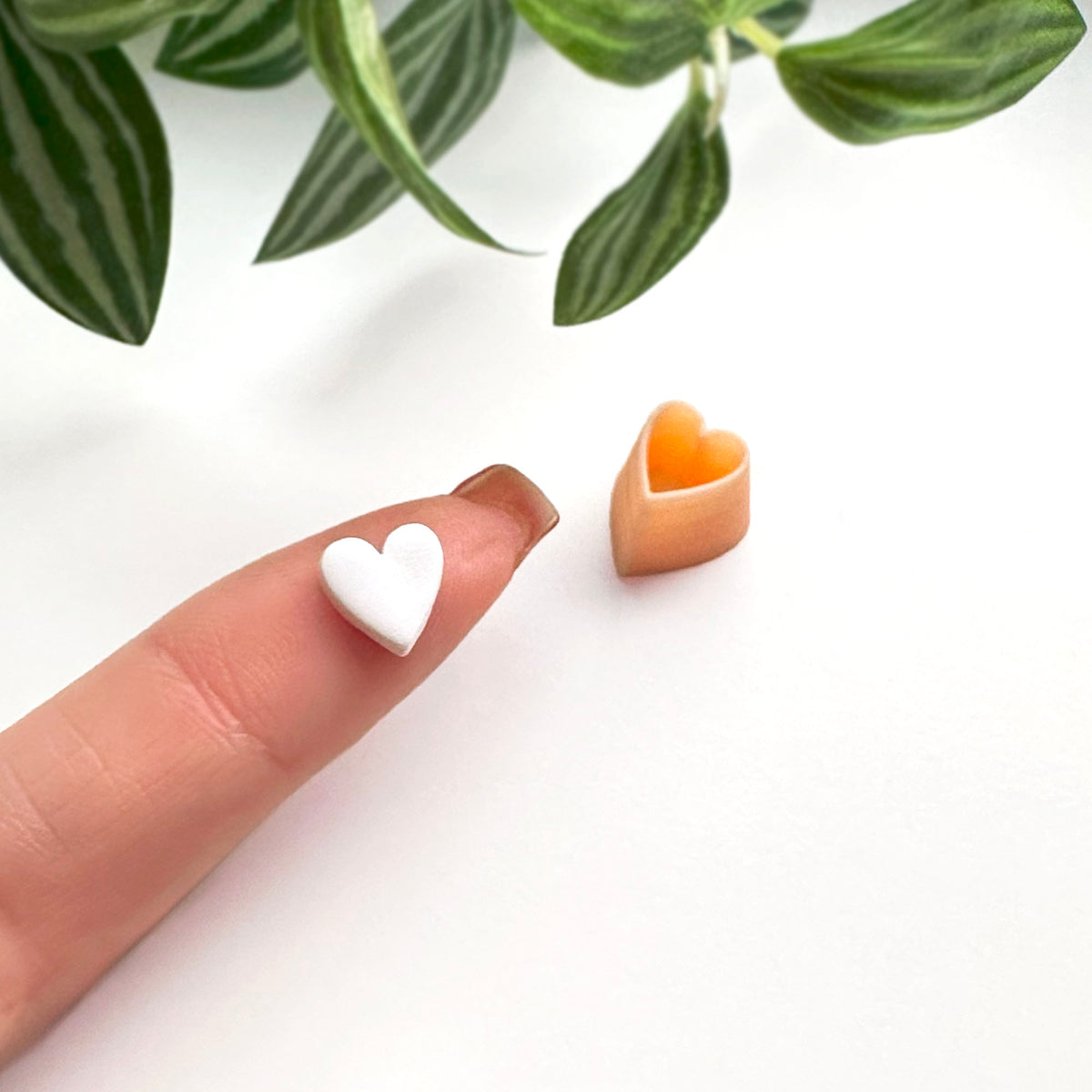 A close-up of a small mini white heart-shaped clay cut out on a finger next to a peach-colored heart-shaped clay cutter, with green plant leaves in the background.