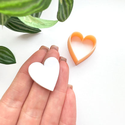 A hand holds a small white heart-shaped clay cut out while an orange heart-shaped cookie clay cutter sits on a white surface, with green leaves in the upper left corner.