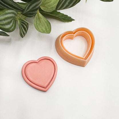 heart-shaped cookie cutter, with pink polymer clay cut out displayed on a white surface with green leaves in the corner.