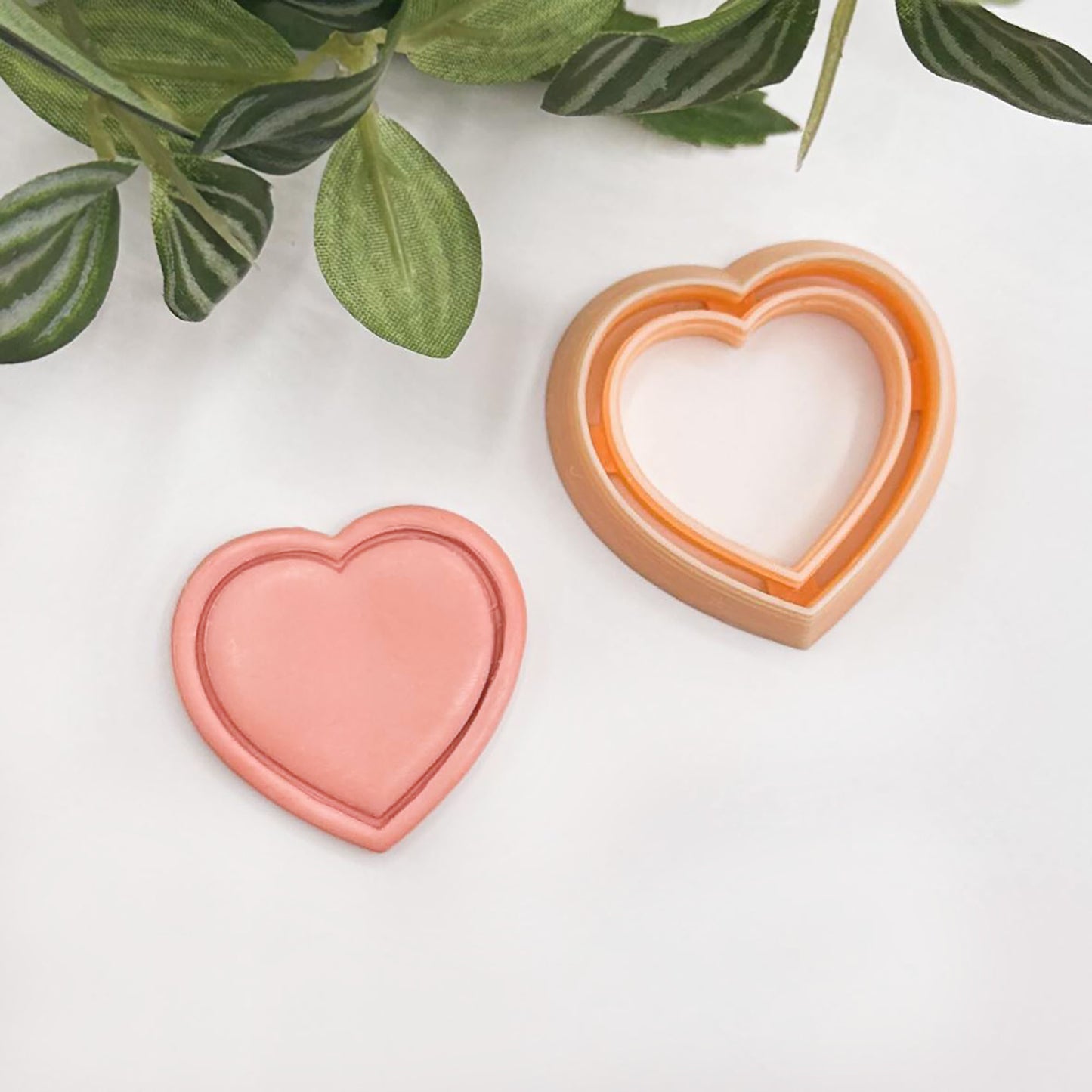 Heart-shaped cookie cutter in peach and orange tones, displayed on a clean white surface with decorative green leaves in the top corner.