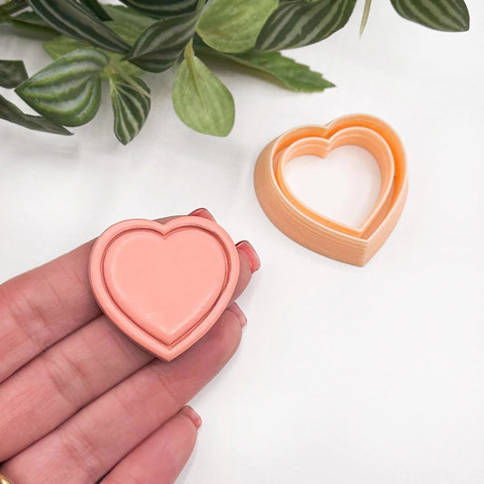 A heart-shaped polymer clay piece in a peach-pink color is held in a person's hand, with the matching peach-colored clay cutter placed on a white surface nearby and green leaves in the upper background.