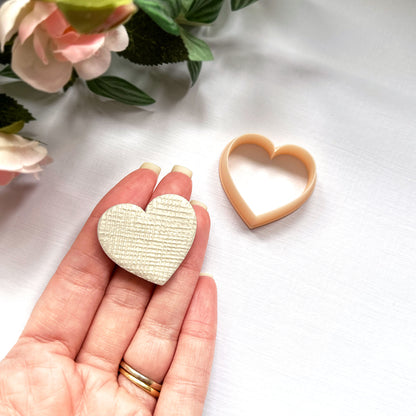 A hand holds a heart-shaped textured cream polymer clay cut-out beside a peach-colored heart-shaped cookie clay cutter on a white surface with soft pink flowers in the background.