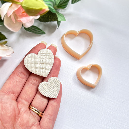A hand holds two small heart-shaped textured cream polymer clay cut-out charms beside two peach-colored heart-shaped cookie clay cutters on a white surface with soft pink flowers in the background.