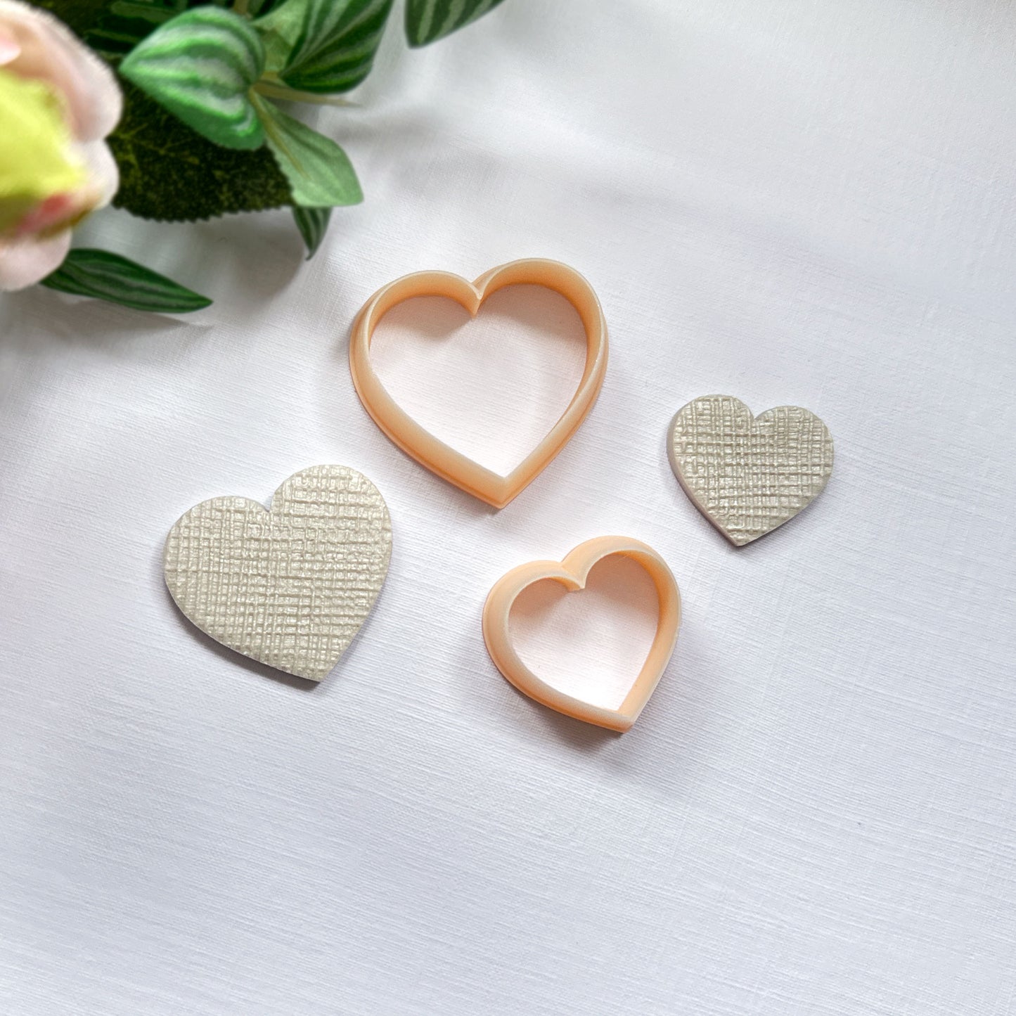 Two heart-shaped textured cream polymer clay cut-out charms beside two peach-colored heart-shaped cookie clay cutters on a white surface with soft pink flowers in the background.