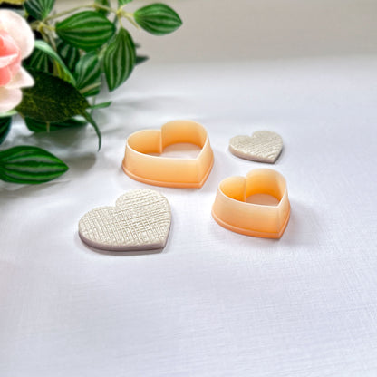Two heart-shaped textured cream polymer clay cut-out charms beside two peach-colored heart-shaped cookie clay cutters on a white surface with soft pink flowers in the background.