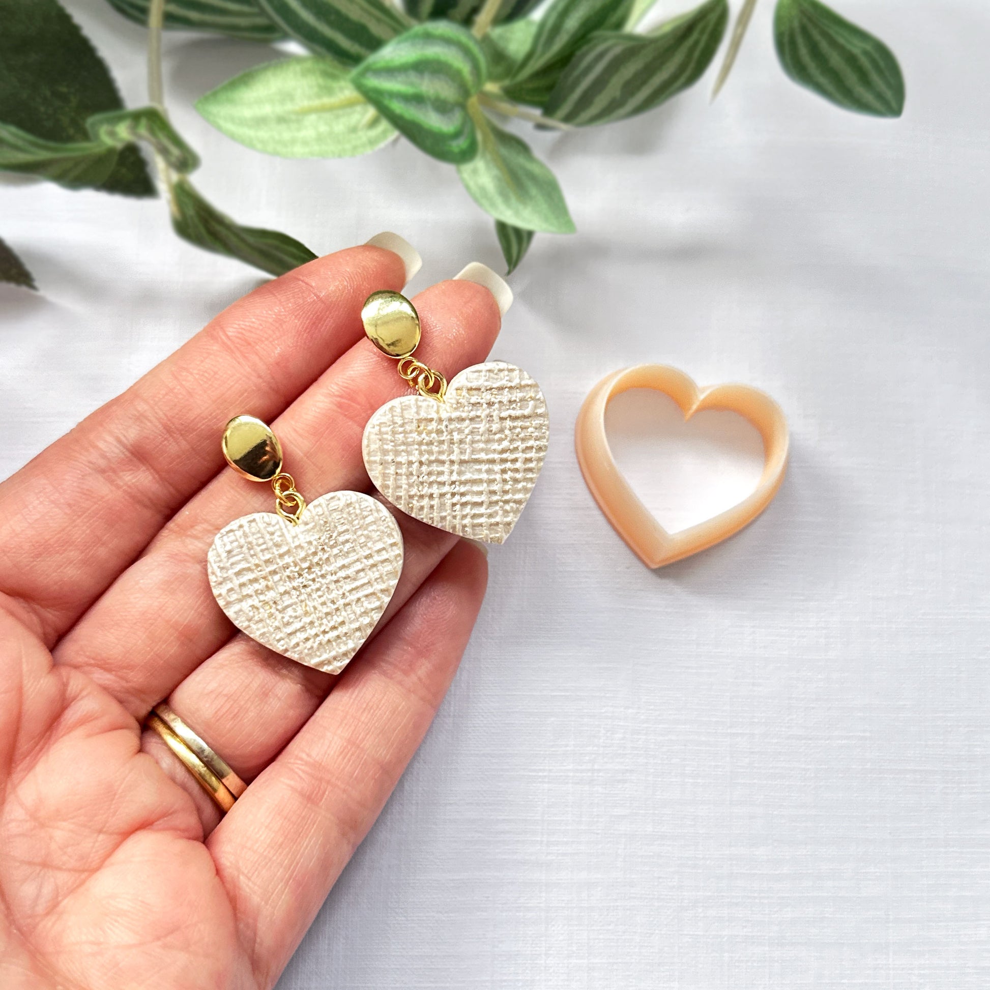 A pair of polymer clay heart-shaped drop earrings with a textured cream face and gold-tone posts, held in a person's hand next to a peach-colored heart-shaped clay cutter on a white fabric background with green leaves.