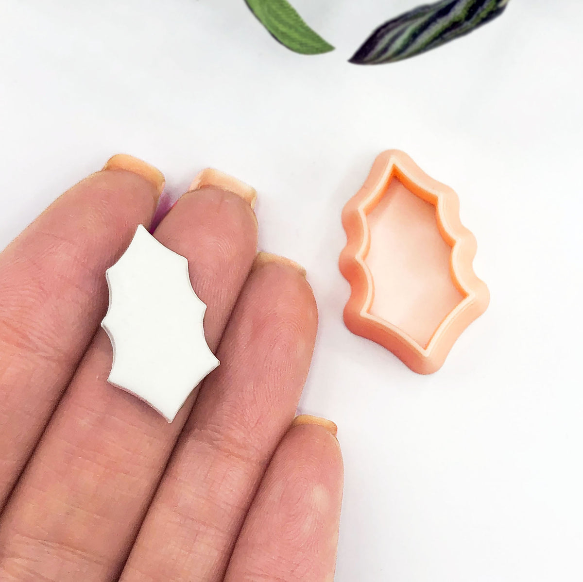 A small white clay cutout held between fingers next to an orange clay cutter on a white surface, with a small plant leaf visible at the top edge.