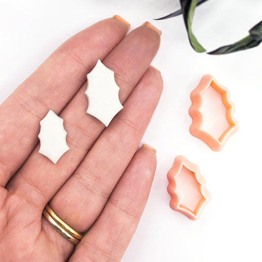 A hand holds two small white holly leaf cut outs, with two peach-colored matching cutters placed beside it on a white background.